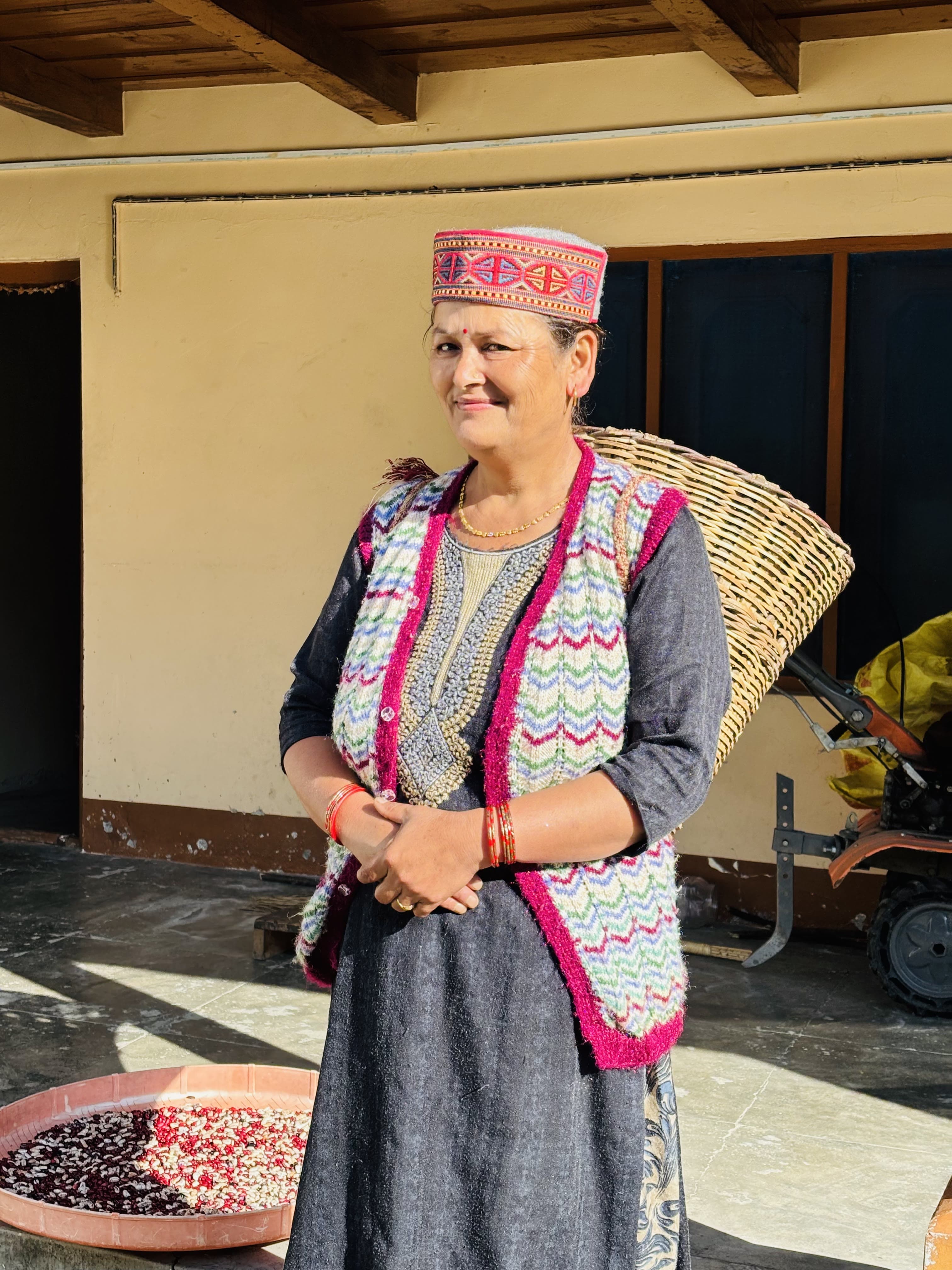 Mummy ji in traditional Himachali dress with a woven farming basket and a tray of drying grain, hand-tiller behind her
